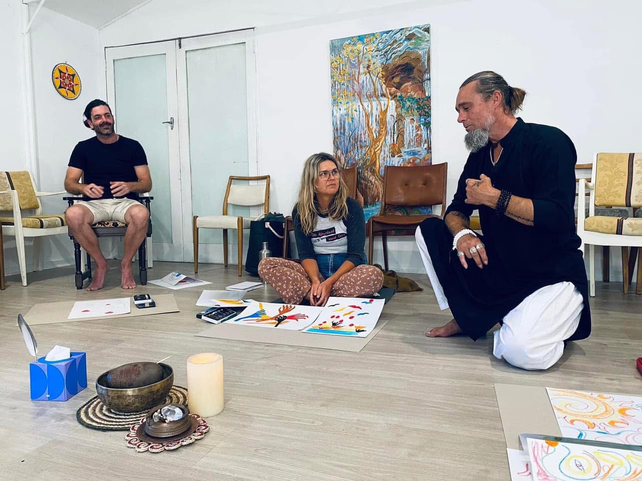 A workshop gathering with drawings and pastels spread on the floor as participants share a conversation.